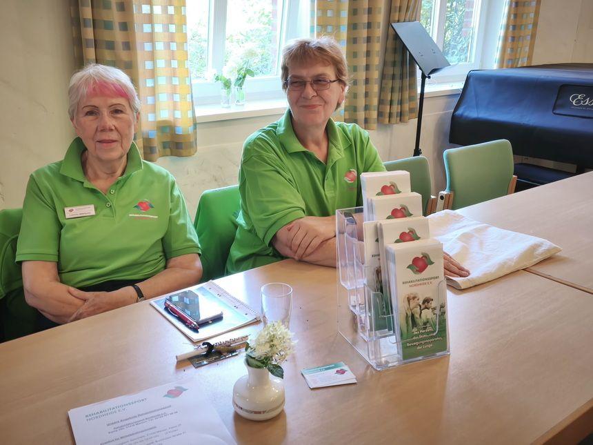 Zwei ältere Frauen in grünen Poloshirts sitzen an einem Tisch mit Prospekten und einem kleinen Blumenstrauß in einer Vase