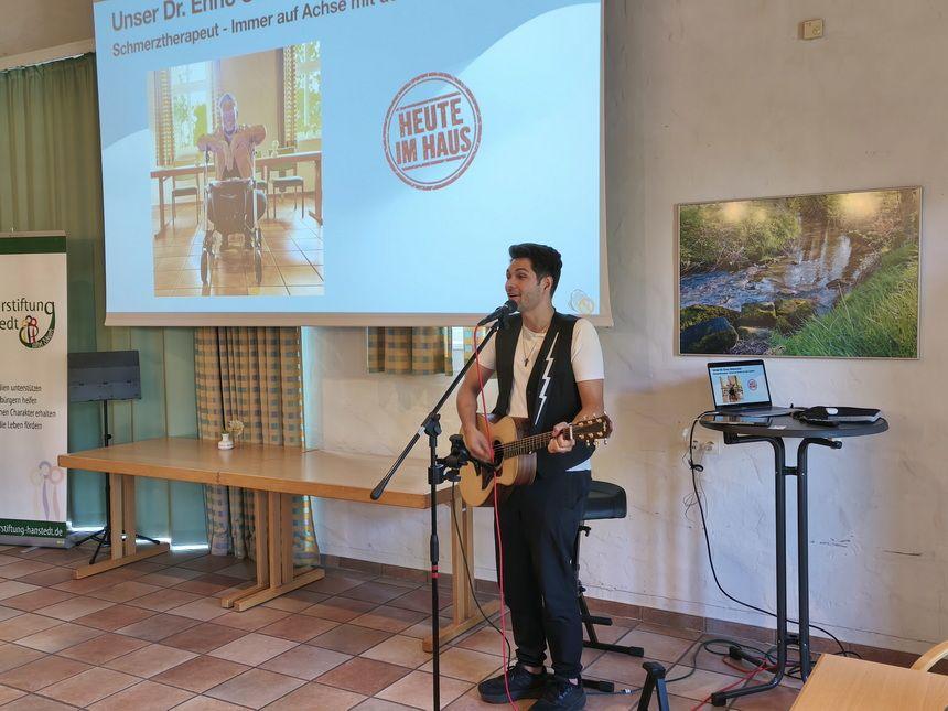 Mann spielt Gitarre und singt vor einer Leinwand mit Präsentationsfolie in einem Raum