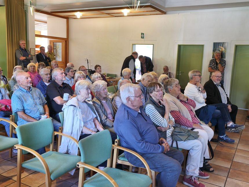 Ältere Menschen sitzen in einem Veranstaltungsraum in Reihen auf Stühlen und hören aufmerksam zu