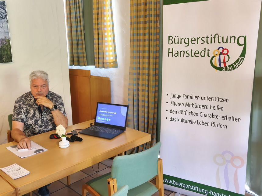 Mann sitzt an einem Tisch neben einem Laptop und einem Banner der Bürgerstiftung Hanstedt