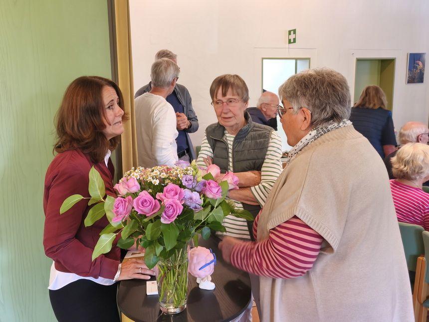 Vier ältere Menschen unterhalten sich um einen Tisch mit einem Blumenstrauß aus rosa Rosen und anderen Blumen in einem hellen Raum.