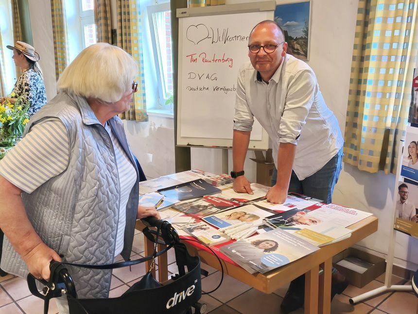 Ältere Frau mit Rollator spricht mit einem Mann an einem Tisch mit Informationsbroschüren in hellem Raum