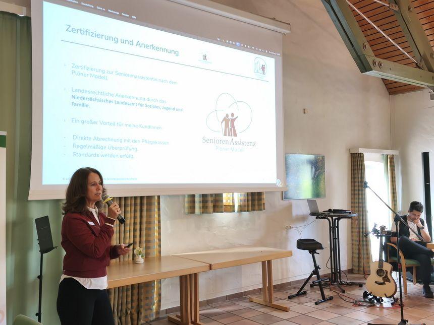 Frau hält eine Präsentation vor einer Projektionswand mit dem Thema SeniorenAssistenz Pionier Modell in einem Saal mit Akustikdecken und Sitzgelegenhe