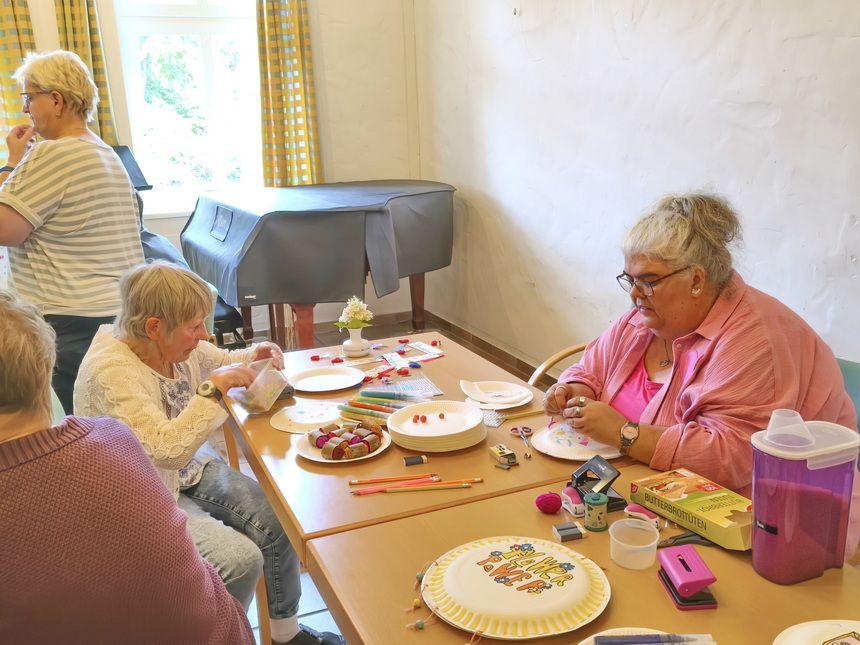 Vier ältere Frauen sitzen an einem Tisch und basteln mit Papier und Farben in einem hellen Raum mit Fenster und Klavier