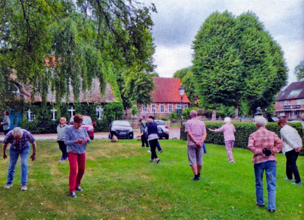 Eine Gruppe von Menschen steht auf einer großen, grünen Wiese vor historischen Gebäuden und Bäumen.